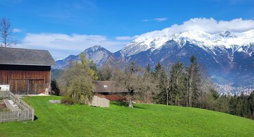 Fotografie: im Vordergrund eine grüne Wiese, links zwei Gebäude einer Hofstelle, im Hintergrund Blick auf die Berge und Innsbruck